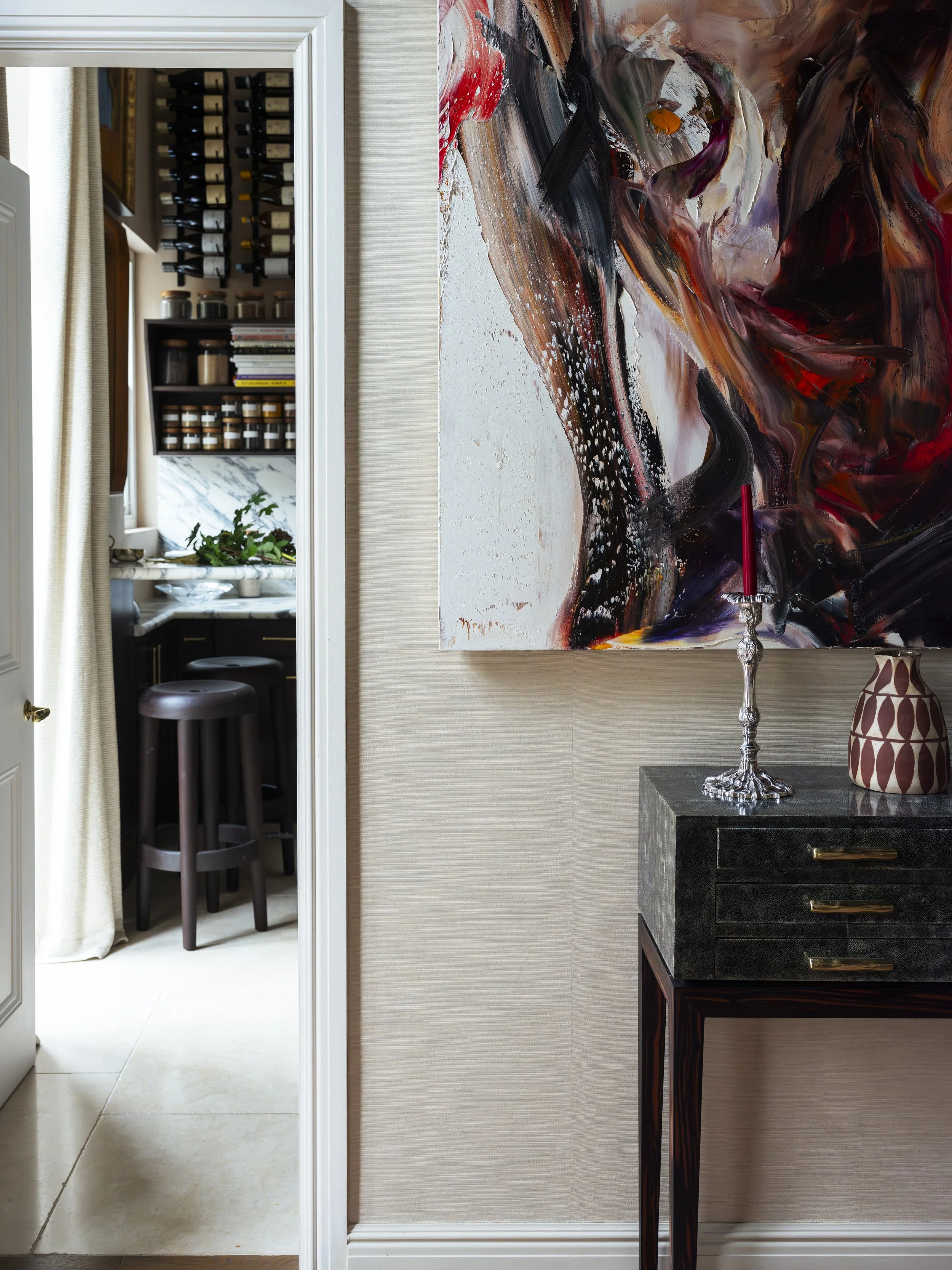 Interior view of a room with a large abstract painting on the wall, a black side table with a pitcher-shaped vase, a silver candlestick with a red candle, and a doorway leading to a kitchen area with shelves of spices and jars, a small marble counter