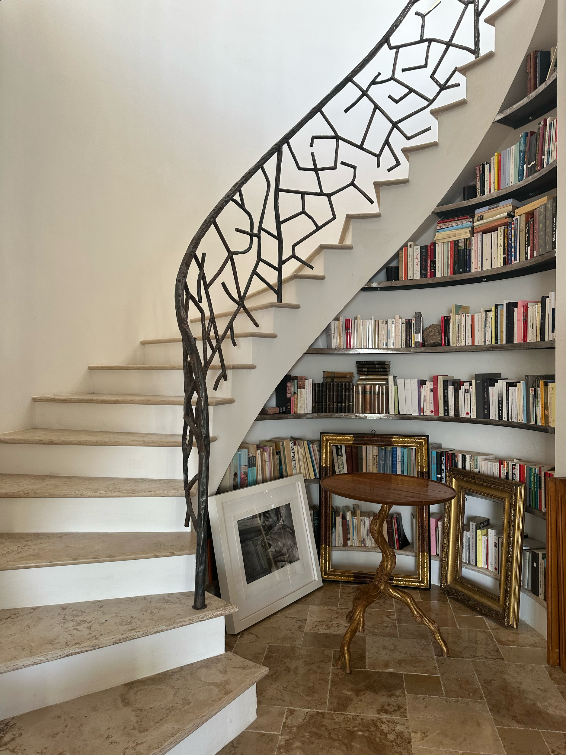 Interior of a home featuring a curved staircase with a decorative black metal railing resembling tree branches, a built-in curved bookshelf filled with books, and a small wooden table with a twisted carved leg, with several empty picture frames and a