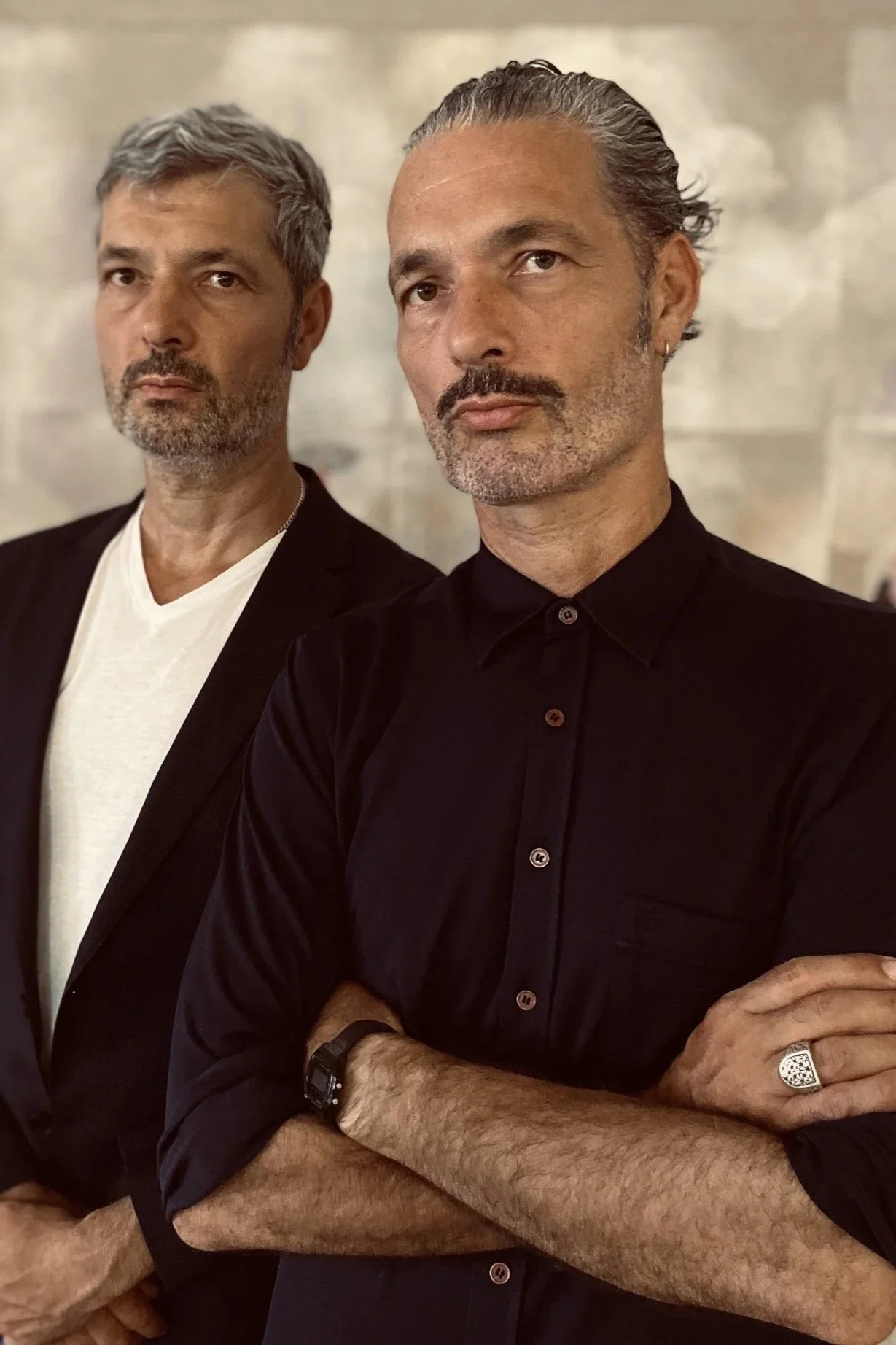 Two men with graying hair and beards standing with arms crossed against a neutral background, looking serious.