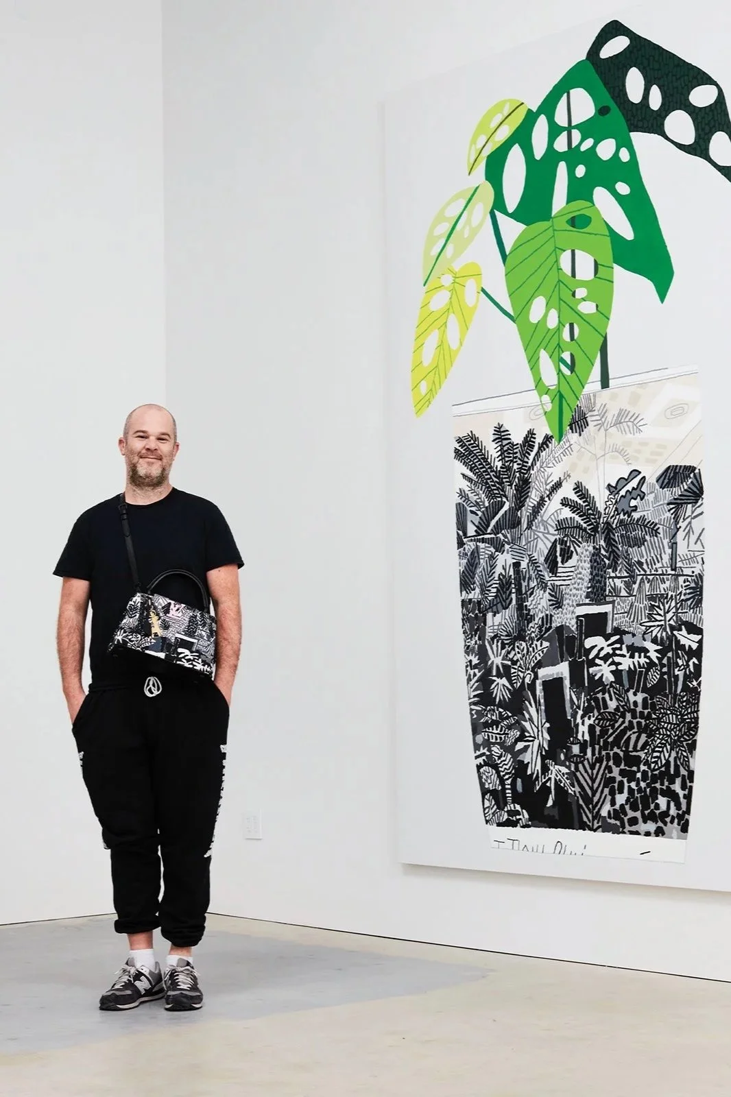 A man standing in an art gallery next to a large black and white tropical jungle print with green leaf illustrations over the top.
