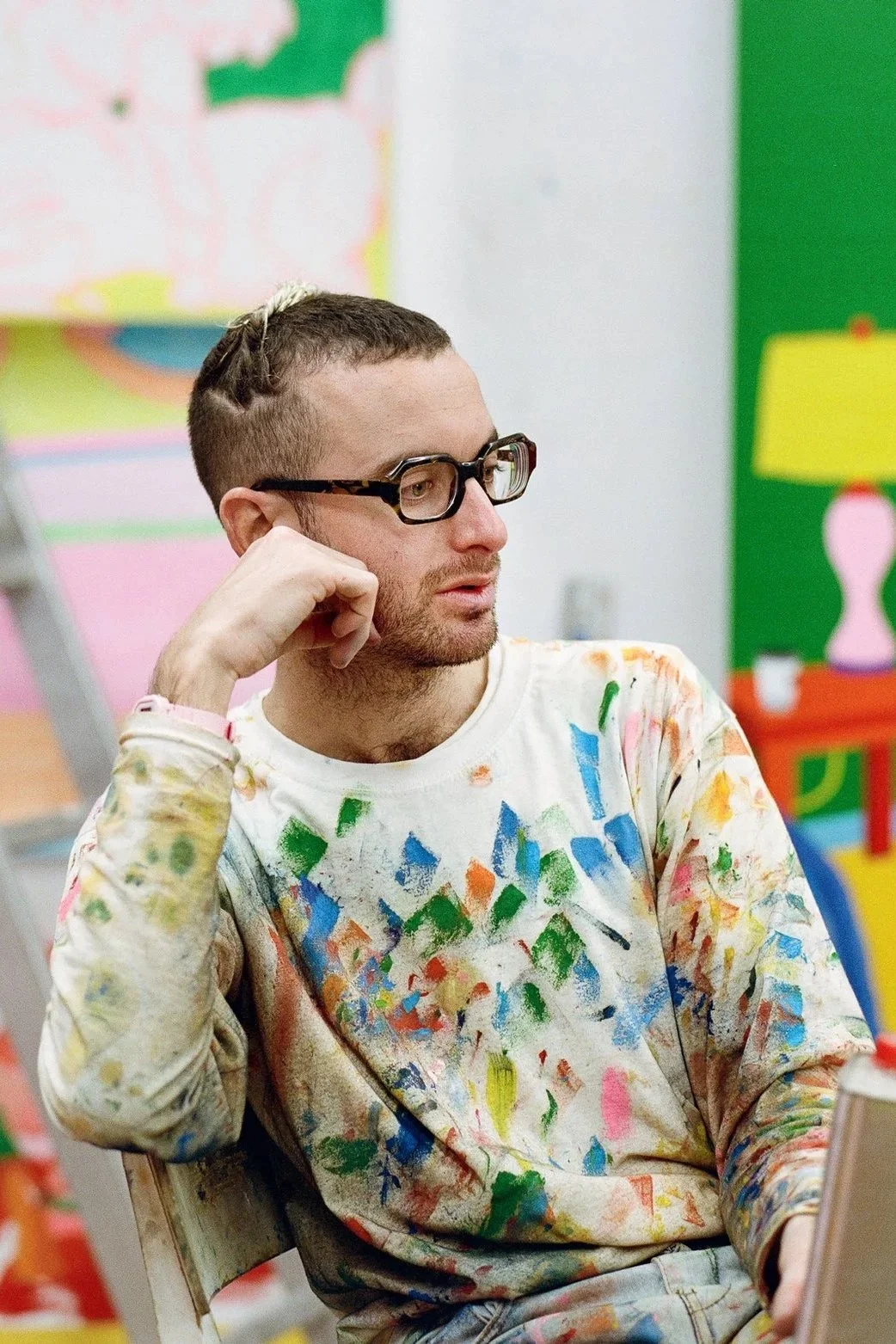 A man wearing glasses and a paint-splattered shirt rests his chin on his hand while sitting in an art classroom.