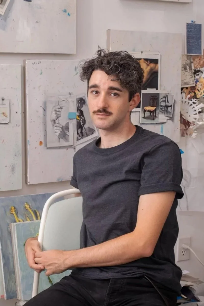 A young man with dark, curly hair and a mustache sitting on a chair in an art studio. The background features various paintings and sketches taped to the wall.