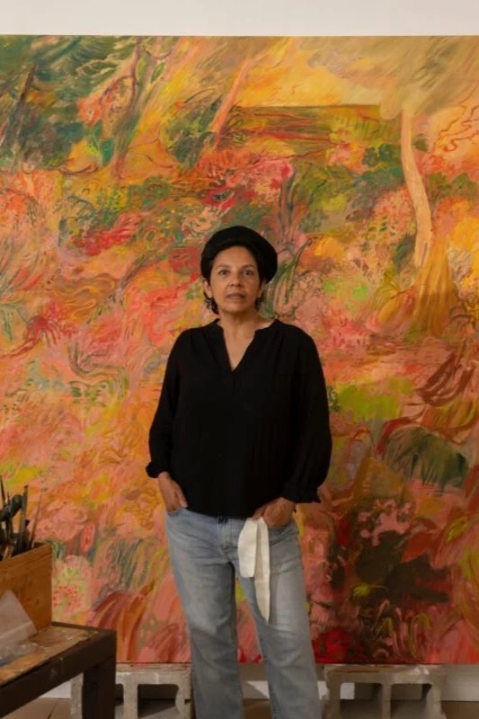 A woman with short dark hair in a black top and light blue jeans stands in front of an abstract, colorful painting.