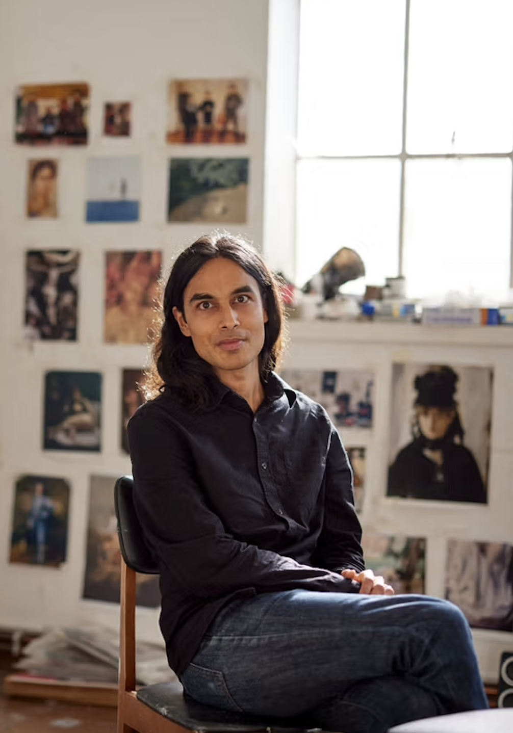 A man with shoulder-length black hair and light skin sits on a chair in a room with a window and a wall decorated with various photographs and artwork.