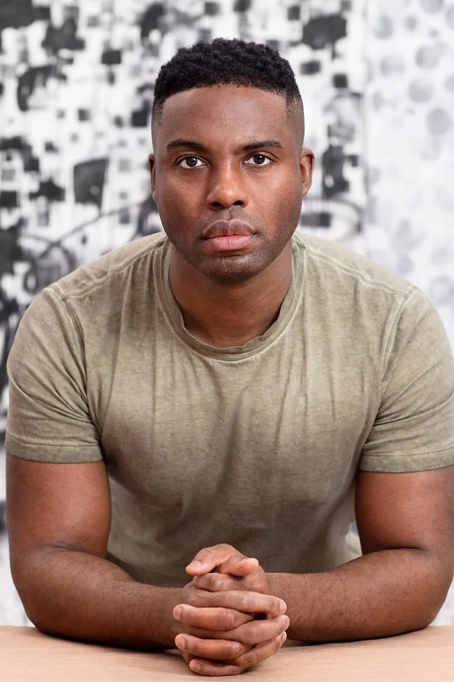 A young man with short black hair and brown skin sits at a table, looking directly into the camera with serious expression, hands clasped in front of him, against a blurred black and white patterned background.