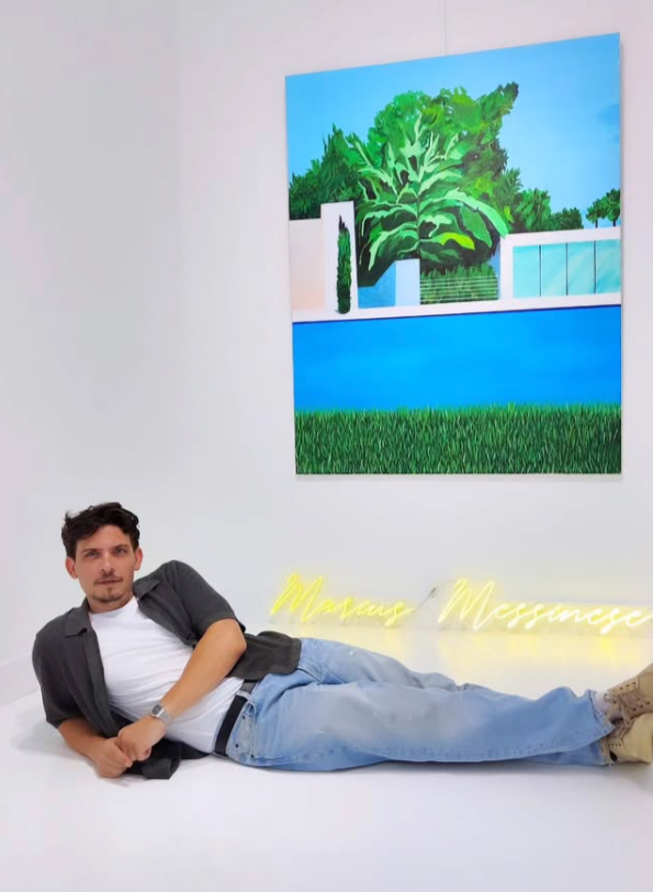 A young man lying on his side in a modern art gallery. Behind him, a large colorful painting of a house with green trees, a blue pool, and a grassy garden. Neon yellow text on the wall reads "Marcus Messange."