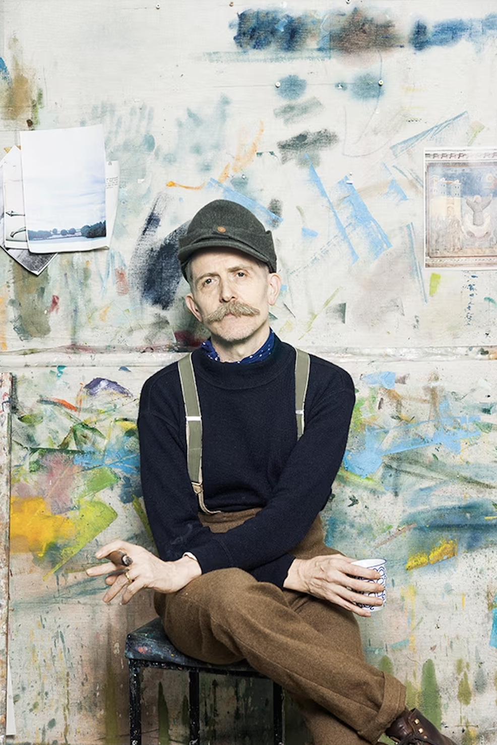 A man with a handlebar mustache and gray hair wearing a dark cap, black sweater, brown pants, and suspenders, sitting on a stool in an art studio surrounded by colorful paint splashes and artwork.
