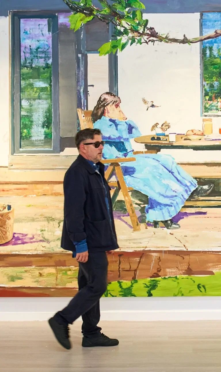 A man with dark hair, sunglasses, wearing a navy jacket, walking past a painting of a woman sitting on a porch, with trees visible through an open window in the background.