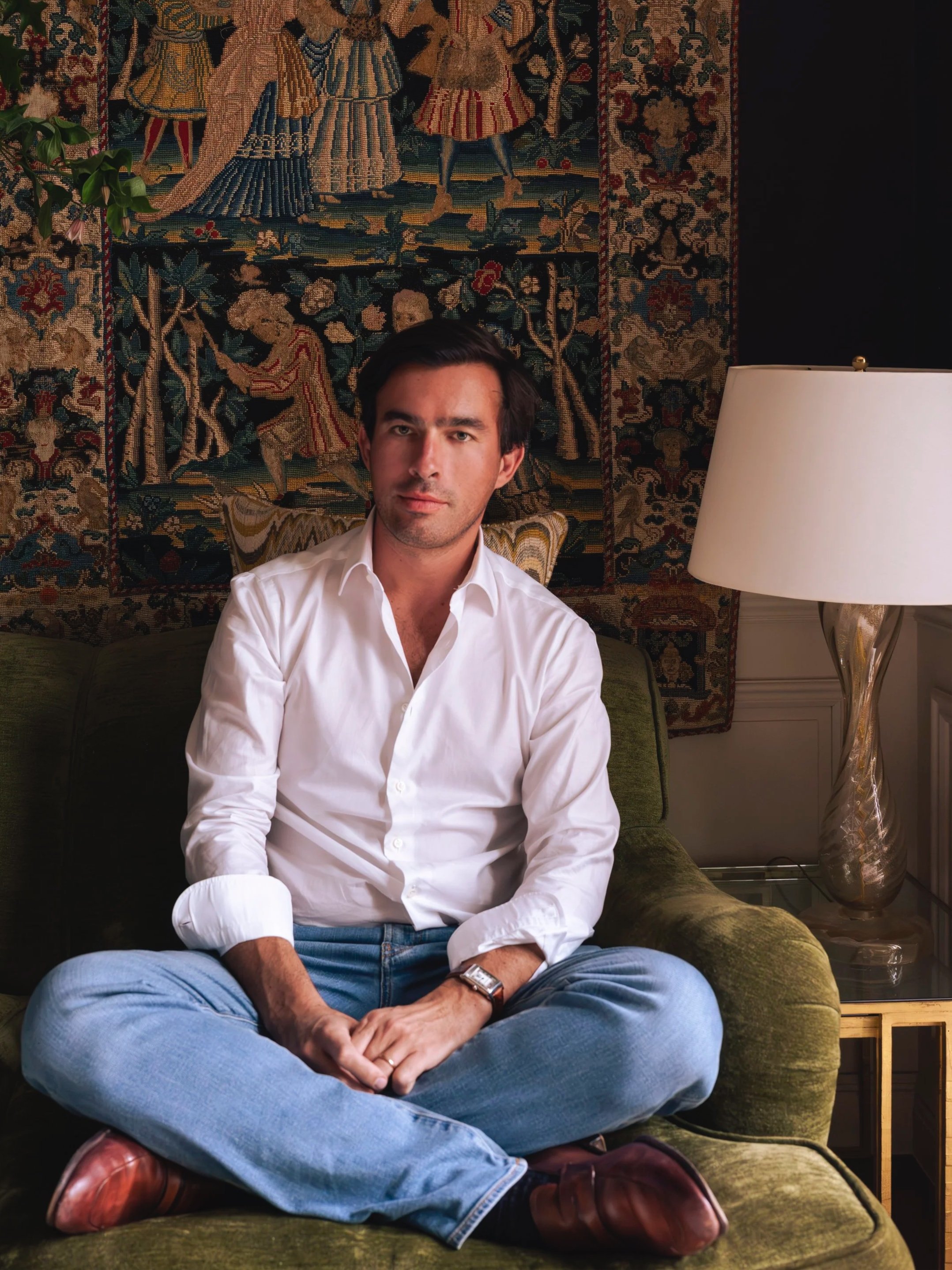 A man sitting cross-legged on a green velvet sofa, wearing a white shirt, blue jeans, and brown shoes, with a beige tapestry hanging behind him and a white lamp on a side table to his right.