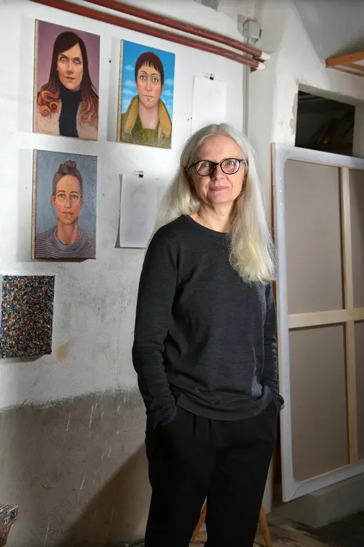 An older woman with long gray hair and glasses standing in an art studio. Behind her are four portrait paintings on the wall, two of women with different hairstyles and expressions. The studio has a white cinder block wall and a large wooden panel leaning against it.