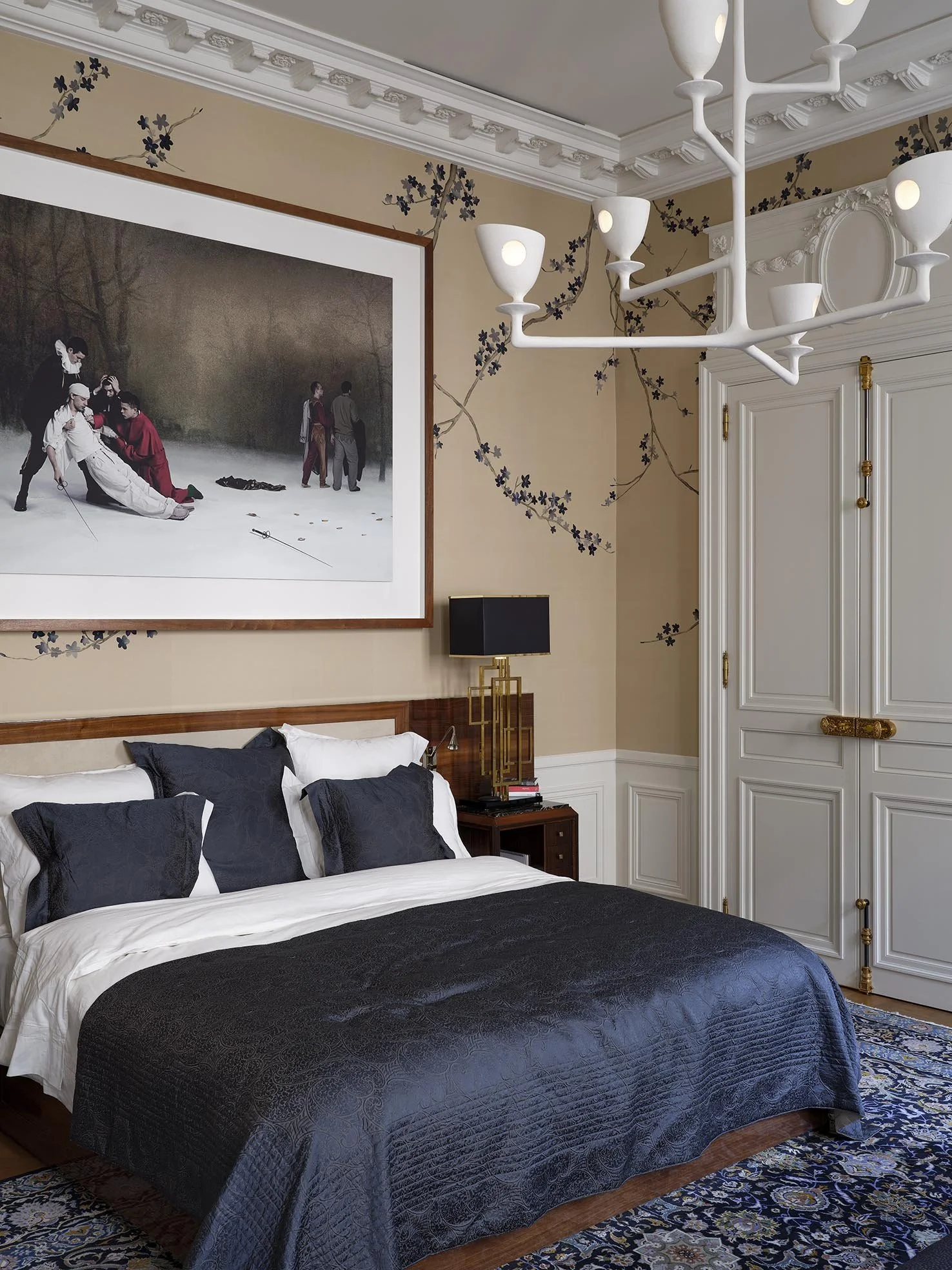 A bedroom with a large bed with black and white bedding and matching pillows. There is a dark wood nightstand with books and a black lamp with a gold base. The wall behind the bed has a large framed painting of a golf scene, and the wallpaper has a f