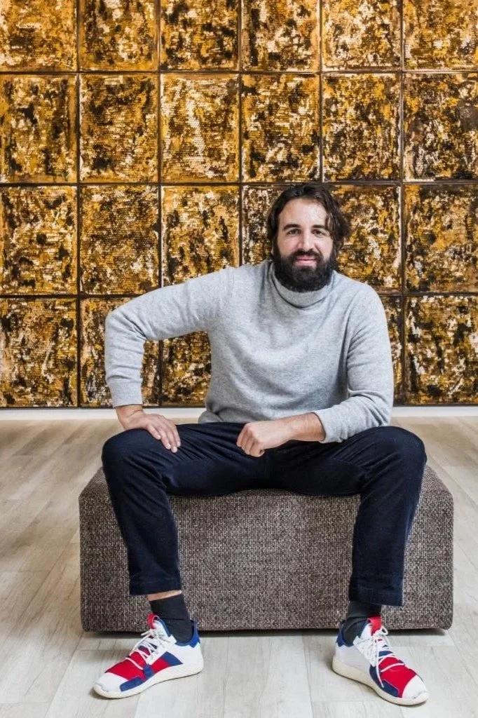 A man with dark hair and a beard is sitting on a brown ottoman in front of a large abstract wall art composed of multiple square panels with mixed gold, black, and white textures. He's wearing a gray sweater, black pants, and colorful sneakers, with a relaxed pose and a slight smile.