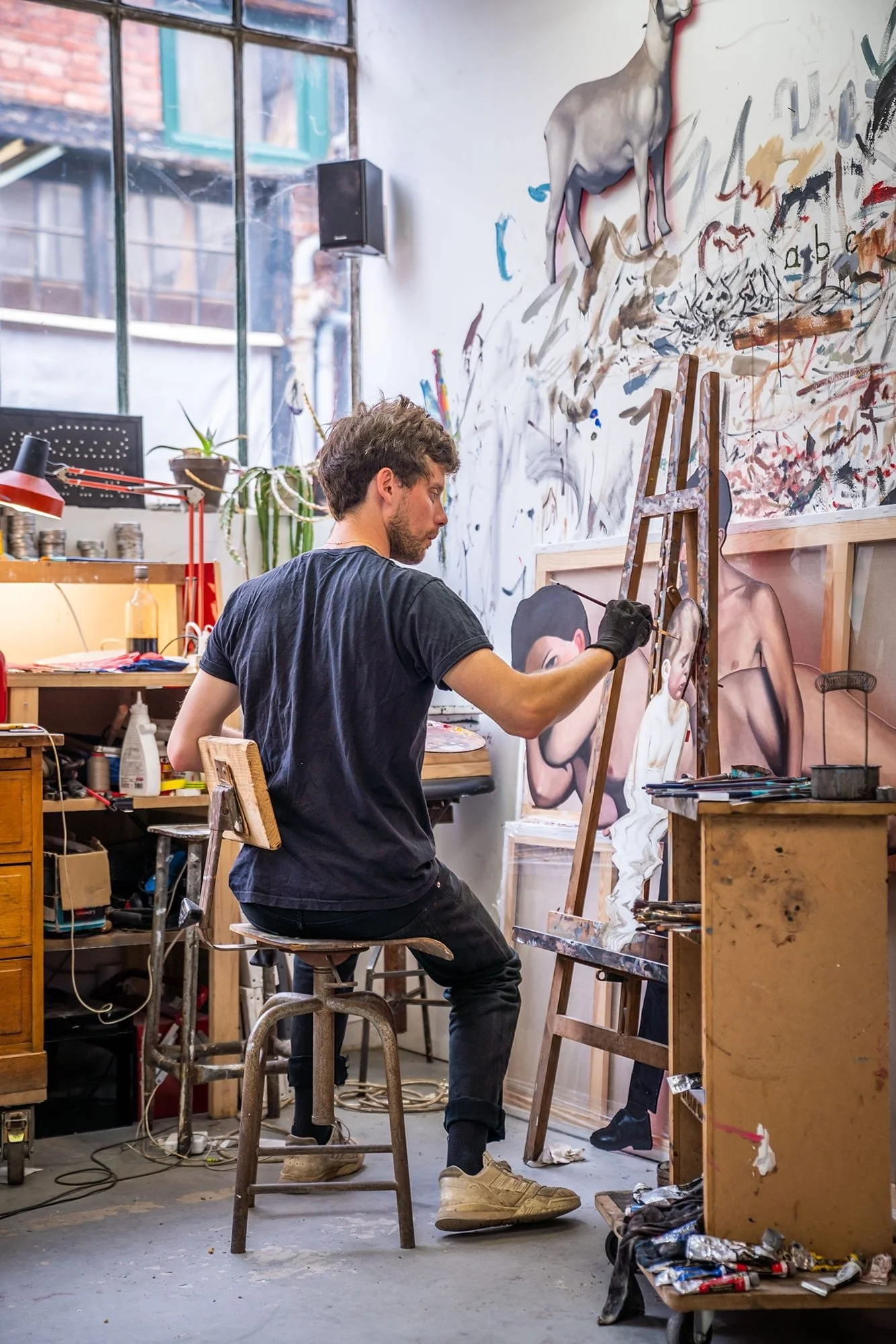 A young man painting on a canvas in an art studio filled with paintings, art supplies, and a large window showing an urban exterior.