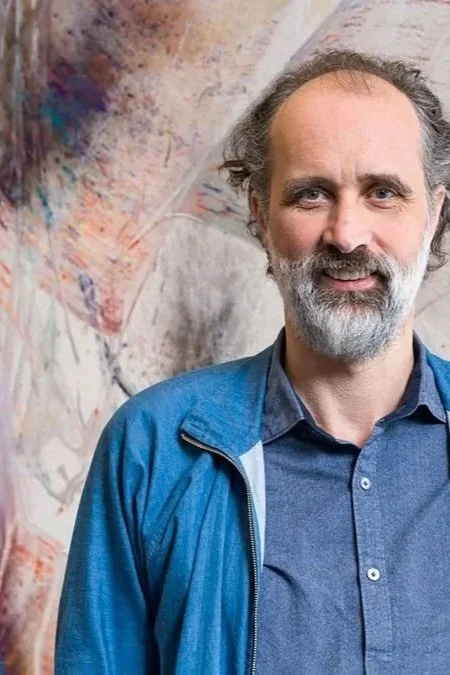 A middle-aged man with a beard and gray hair, wearing a blue jacket over a blue shirt, standing in front of an abstract colorful painting.