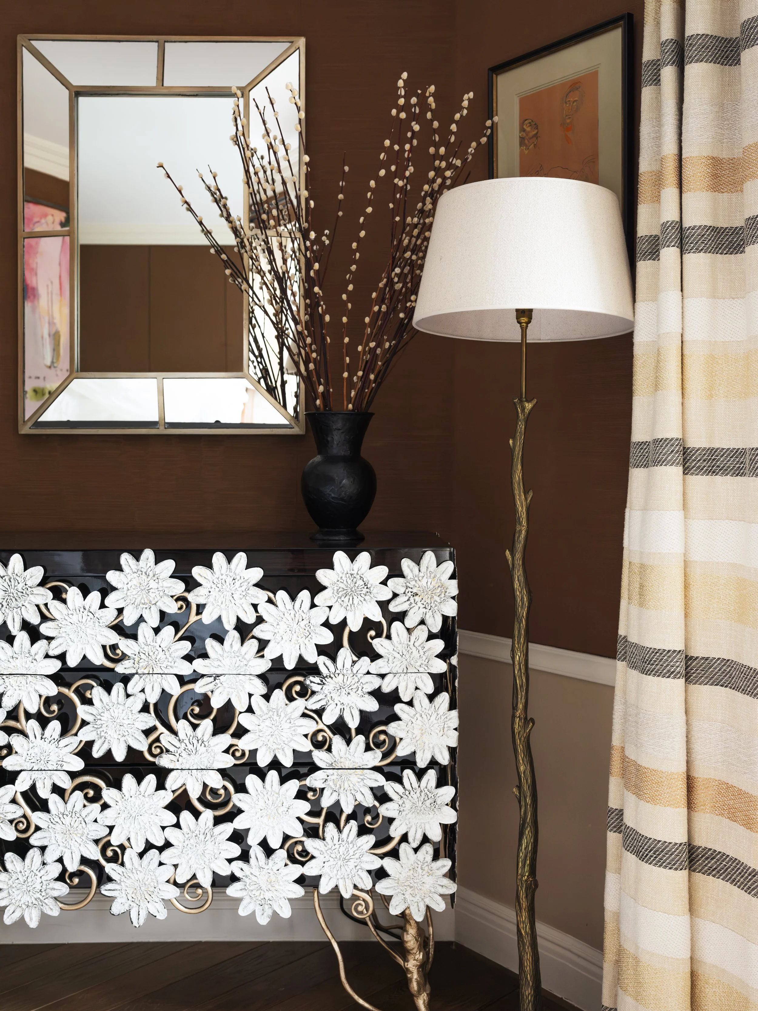 Interior corner with a mirror, a black vase with pussy willow branches, a decorative white floral-patterned console, a standing lamp with a branch-like gold base, and a curtain with horizontal striped pattern.