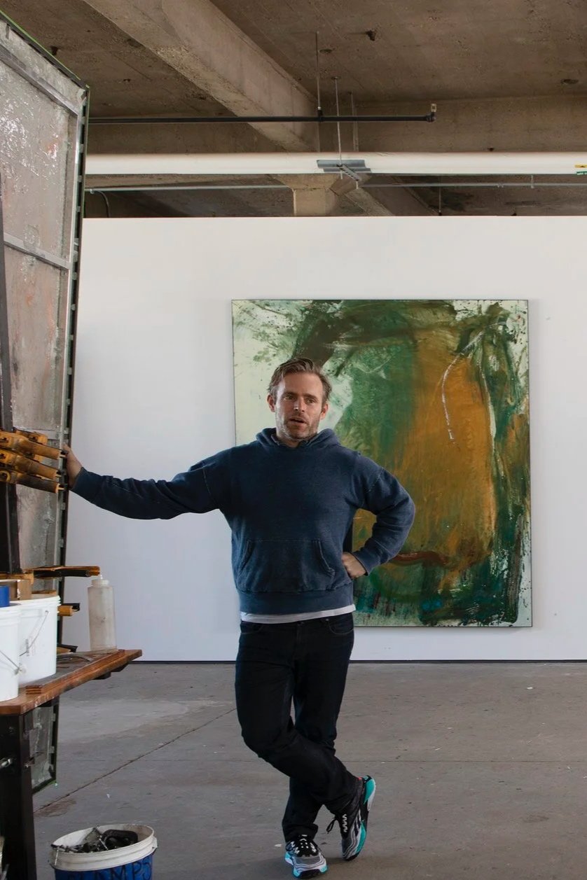 A man in a dark hoodie and black pants standing in an art studio or gallery, with one arm resting on a shelf and the other hand on his waist. Behind him, there is a large abstract painting with green, yellow, and brown colors on a white wall. The studio has an unfinished ceiling with exposed concrete and pipes.