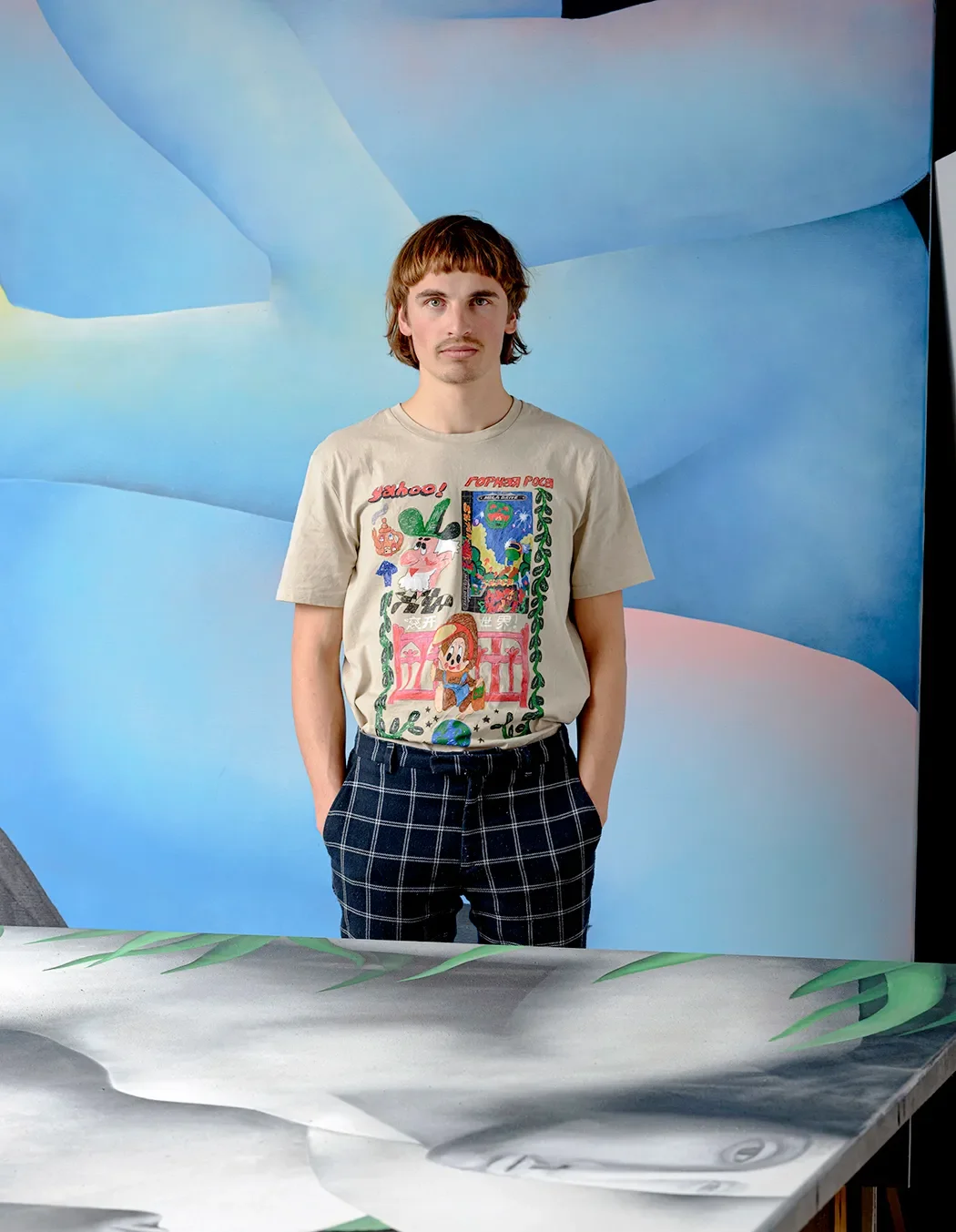 A young man with brown hair and a mustache wearing a graphic T-shirt featuring cartoon characters and dark plaid pants, standing in front of a blue abstract background.
