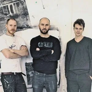Three men standing indoors with arms crossed, casual clothing, artwork on the wall behind them.