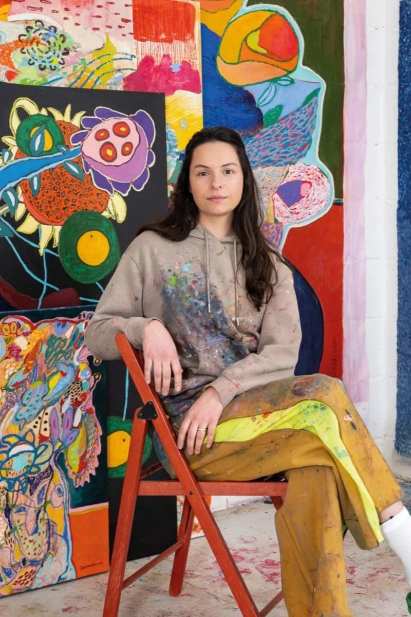 A young woman with long dark hair sitting on a red wooden chair in front of colorful abstract paintings in an art studio. She is wearing a paint-splattered hoodie and pants.