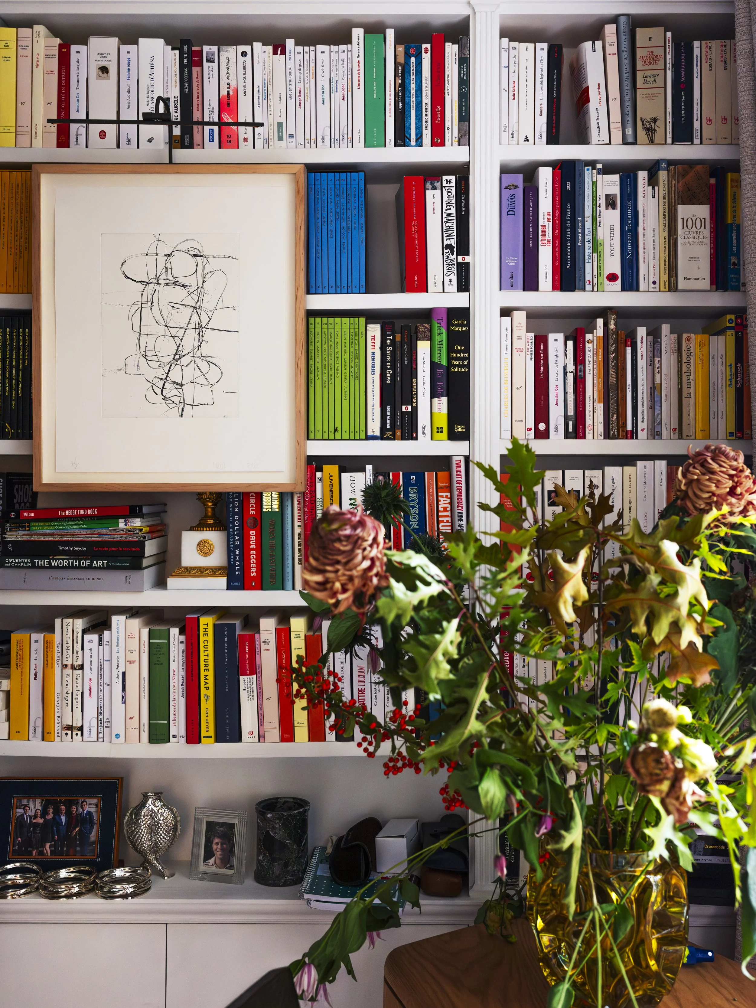 A white bookshelf filled with colorful books, a framed abstract line drawing, a small altar with a gold statue, a photo, and silver decorative objects. In front, a large yellow vase with a bouquet of mixed flowers and foliage.