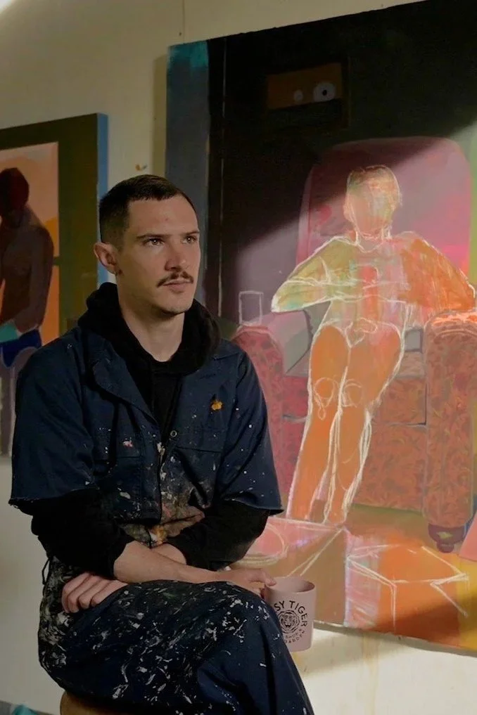 A man with a mustache and dark hair, wearing a black hoodie and paint-splattered paint overalls, sitting on a stool in front of a brightly colored painting of a person playing a guitar on a chair.