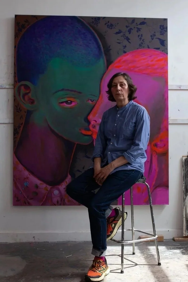 A woman sitting on a stool in front of a large, colorful painting of two abstract faces with exaggerated features in an art studio.