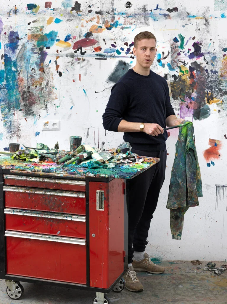 Young man standing in an artist's studio, holding a paintbrush and palette while looking towards the camera. The background features a wall with colorful paint splatters and a worktable covered in art supplies and paint stains.