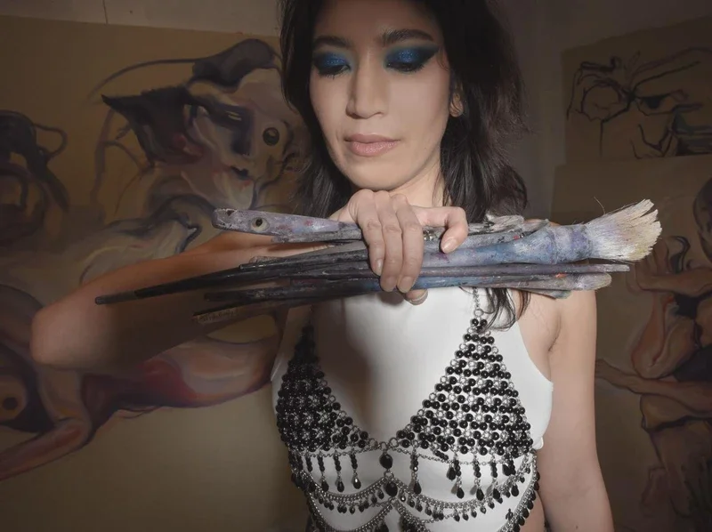 A woman with dark hair and blue eye makeup holds a stack of paintbrushes resembling fish, in front of a colorful mural featuring fantasy creatures.