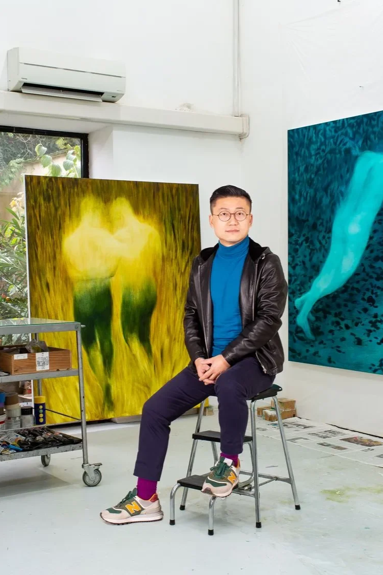 A man sitting on a stepladder in an art studio with two large abstract paintings in the background, one green and yellow and the other blue with a white figure.