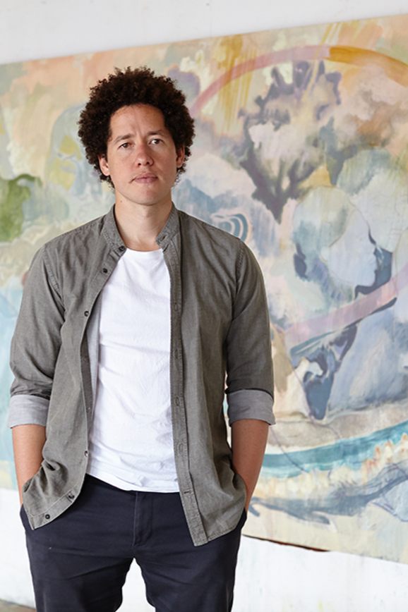 A man with curly hair standing in front of an abstract artwork on the wall.