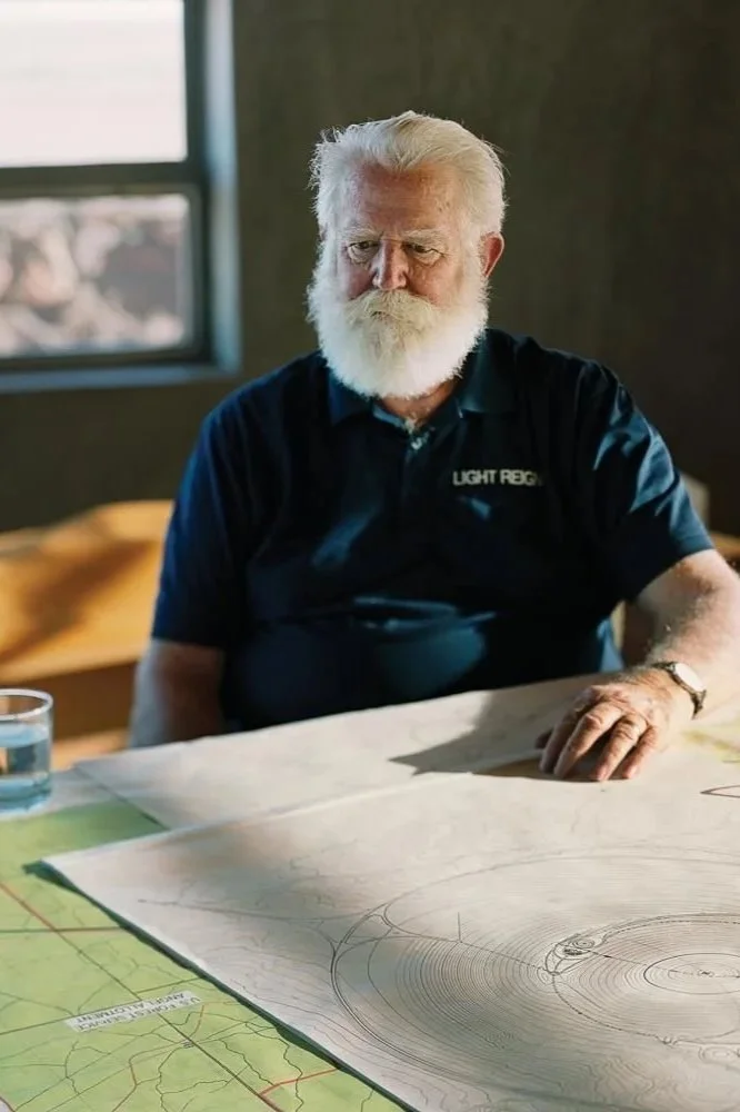 An elderly man with white hair and beard, wearing a navy blue polo shirt with 'Light Reg' on it, sits at a table with a large map and drawings. There is a glass of water on the table and a window in the background.