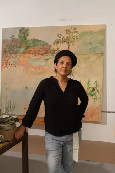A woman standing in front of an abstract landscape painting with trees, water, and sky, in an art studio or gallery.