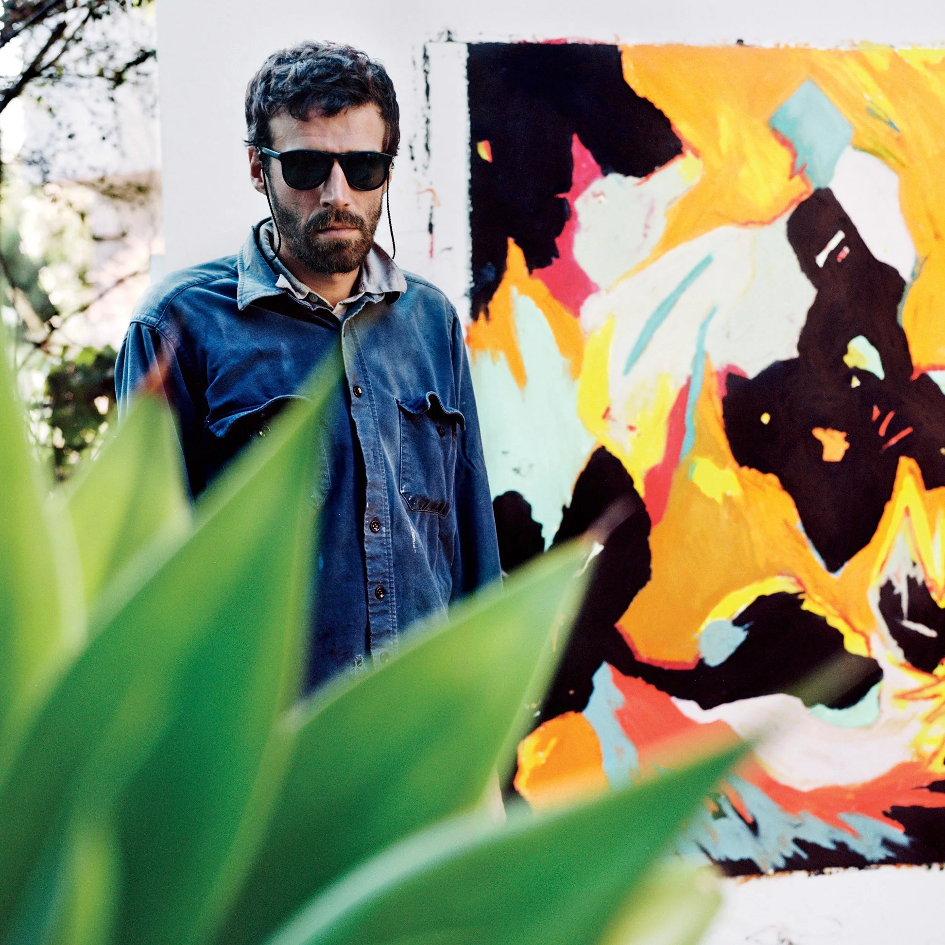 A man with dark hair, beard, and sunglasses standing next to an abstract colorful painting, with green plant leaves in the foreground.