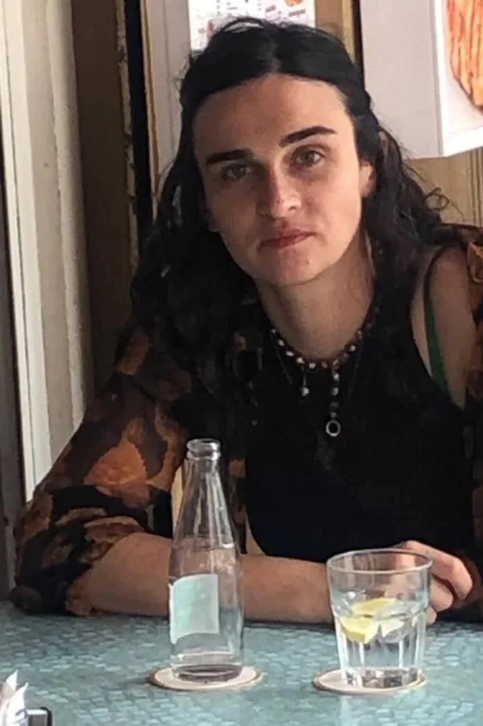 A woman with dark hair and fair skin sitting at a table, with a glass of water with lemon and an empty glass bottle in front of her. She is wearing a dark top with a floral pattern and a beaded necklace, in an indoor setting.