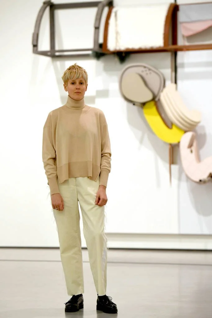 A person with short blonde hair wearing a beige turtleneck sweater and cream-colored pants, standing indoors with abstract wall art in the background.