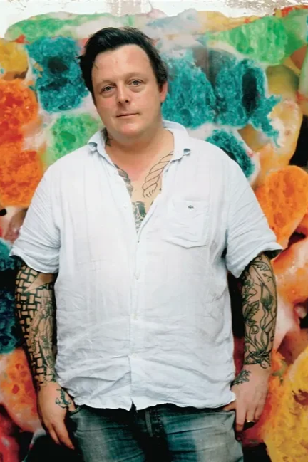 Person with dark hair, wearing a white button-up shirt with rolled sleeves, tattoos visible on forearms and chest, standing in front of a colorful, abstract background.