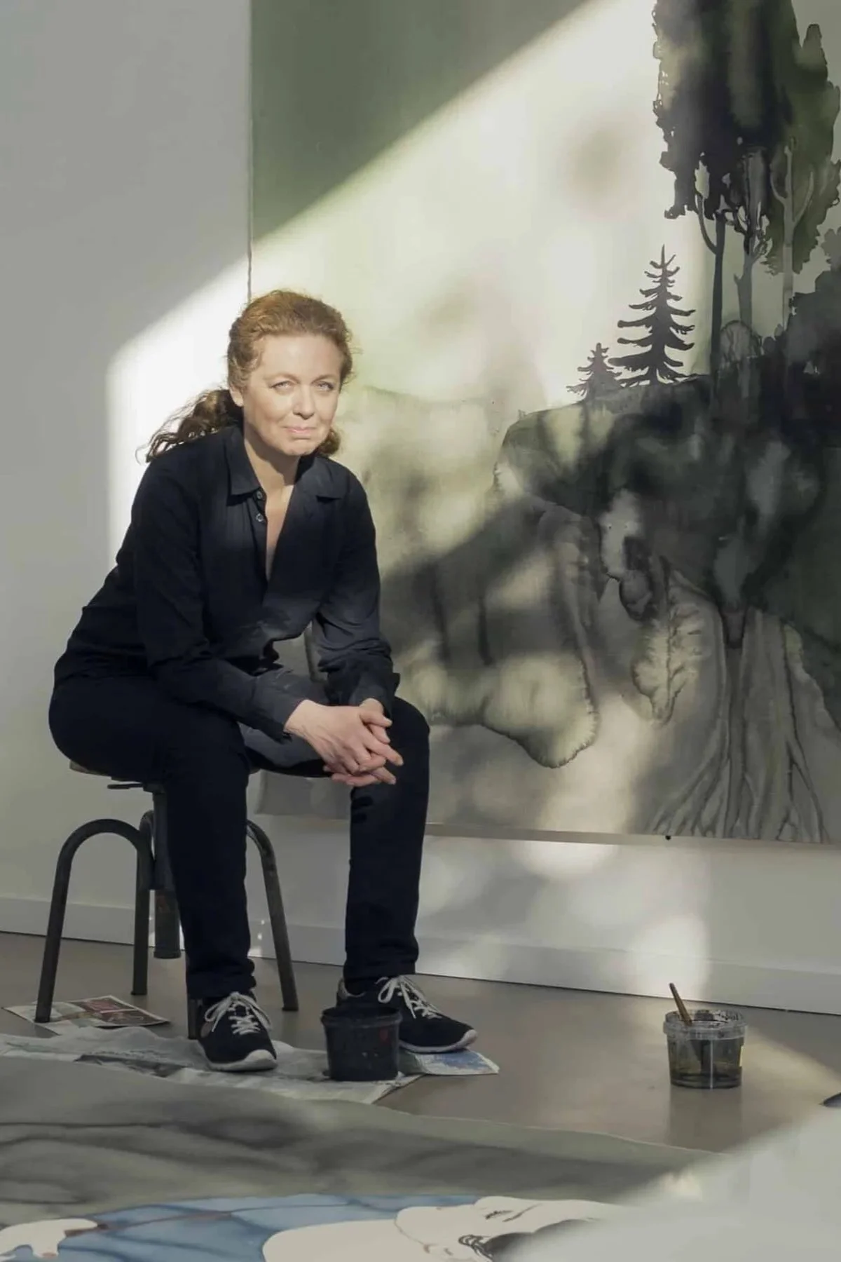 A woman with reddish hair wearing a black shirt and black pants, sitting on a black stool, with art supplies and paper on the floor around her, in front of a large canvas displaying a monochrome landscape painting with trees and mountains.