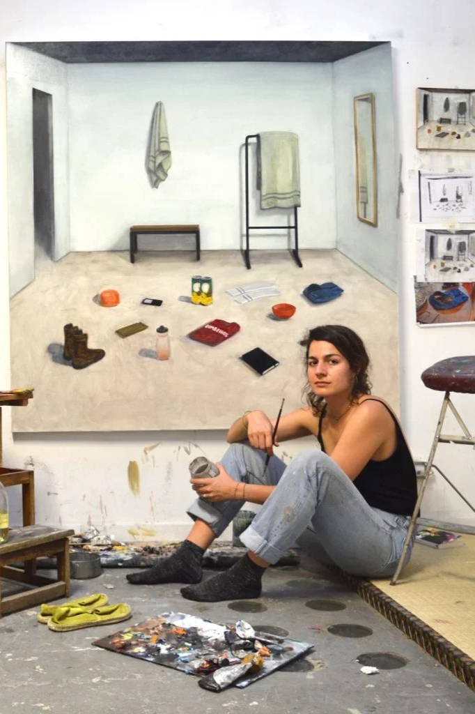 Young woman sitting on the floor in an artist's studio, holding a jar with paint, surrounded by painting supplies, in front of a large, realistic painting of a minimalist room with a towel, shelf, and various objects.