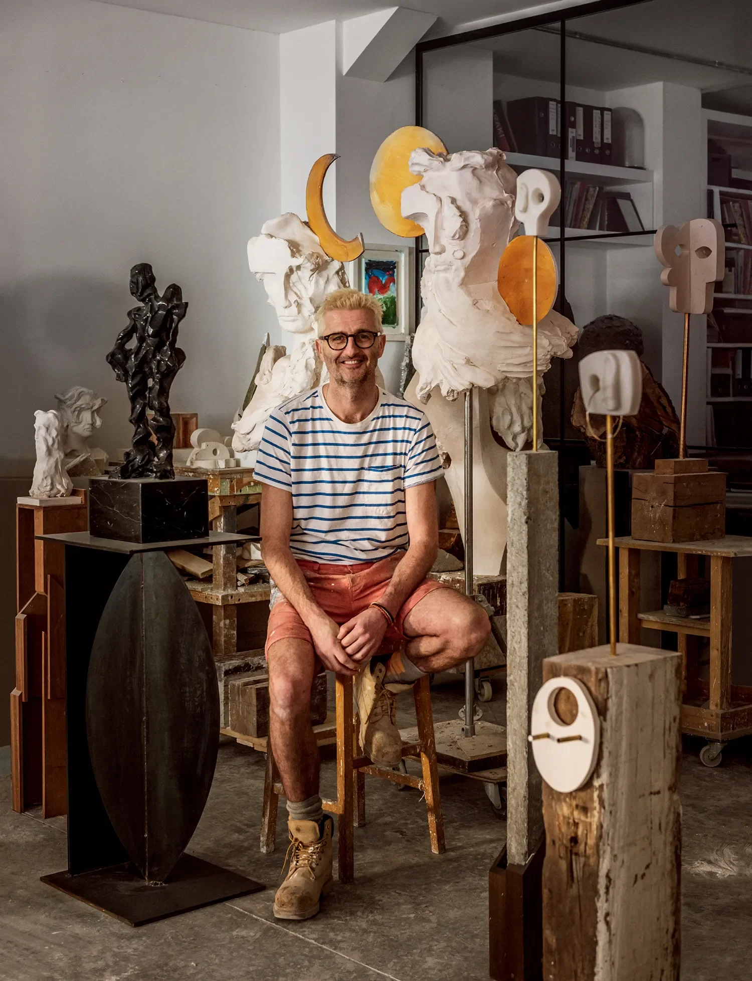 An artist sitting on a wooden stool surrounded by various sculptures and art pieces in a studio, including busts, abstract forms, and geometric pieces.