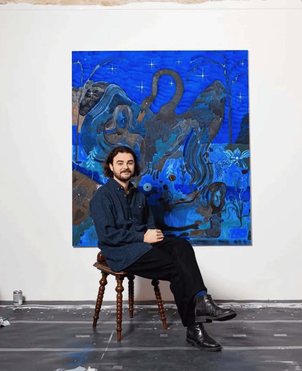 A man with dark hair and beard, wearing a dark blue shirt and black pants, seated on a wooden stool, in front of a large blue abstract painting with swirling shapes and star-like details, in an art gallery.