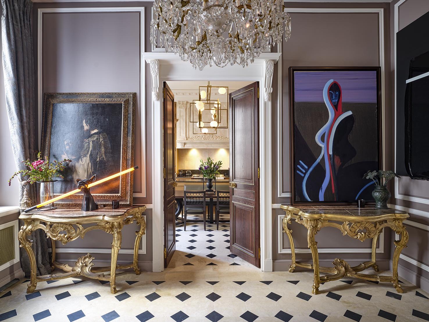 Elegant interior with ornate gold consoles, large modern artwork, a grand chandelier, and a view into a room with a dining table, chandelier, and classic decor.