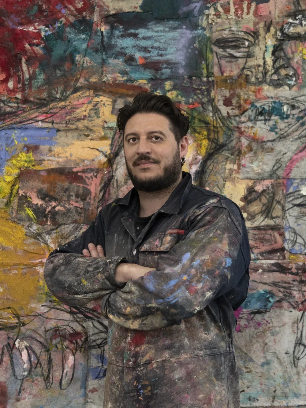 A man with dark hair and a beard, wearing a painted smock, standing with arms crossed in front of a colorful, abstract art piece.