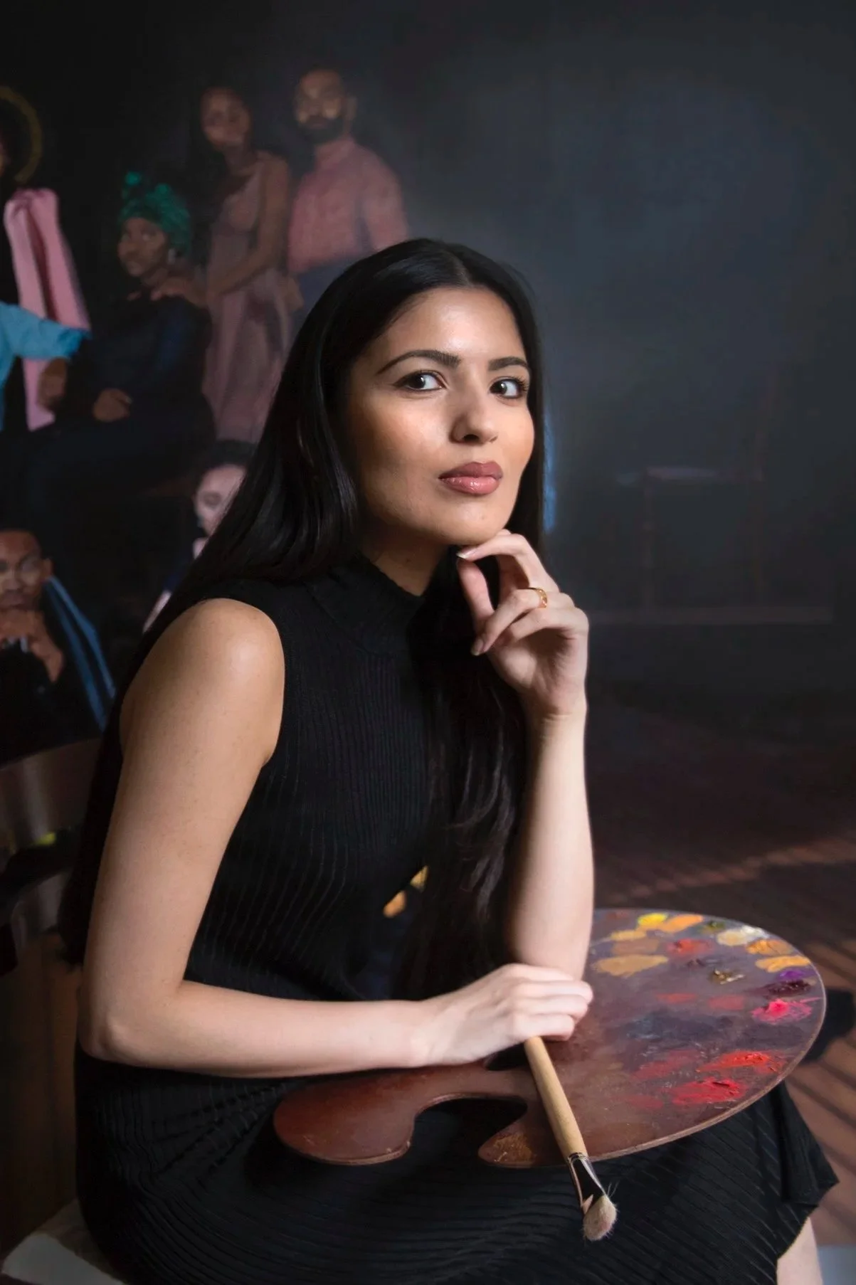 A woman with long black hair and a black sleeveless top sitting at a table holding an artist's palette and brush, with a mural of diverse people in the background.