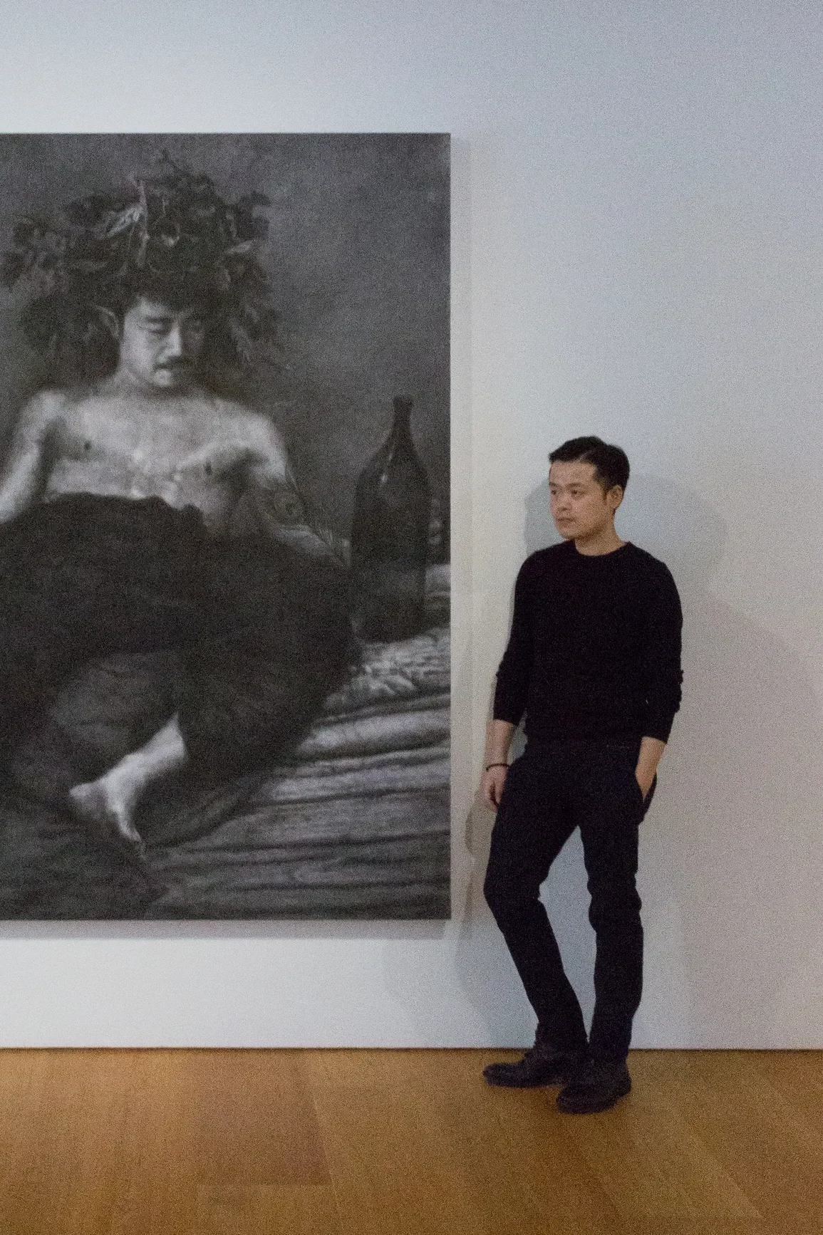 A man stands next to a large black-and-white photograph of a woman with a headdress made of leaves. The woman in the photograph is sitting on a bed with a bottle nearby.