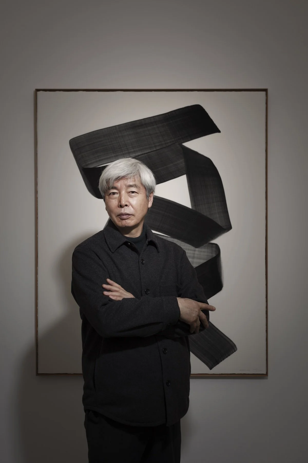 A man with gray hair wearing a black button-up shirt standing with crossed arms in front of an abstract black and gray artwork on a white wall.
