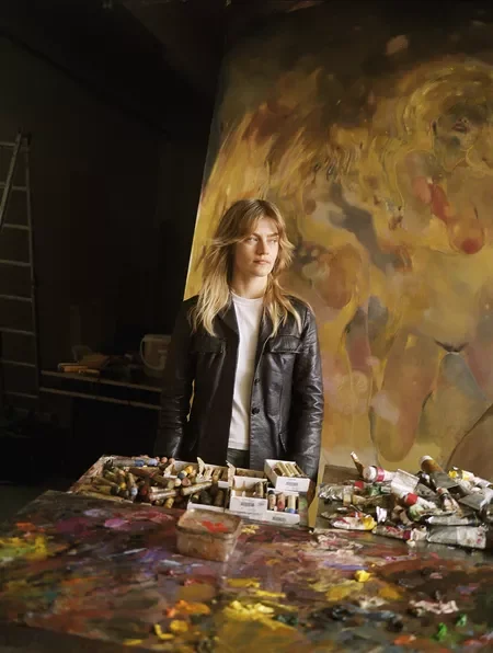 A young woman with blonde hair wearing a black leather jacket stands in an art studio behind a table covered with paint supplies, with an abstract colorful painting on the wall behind her.
