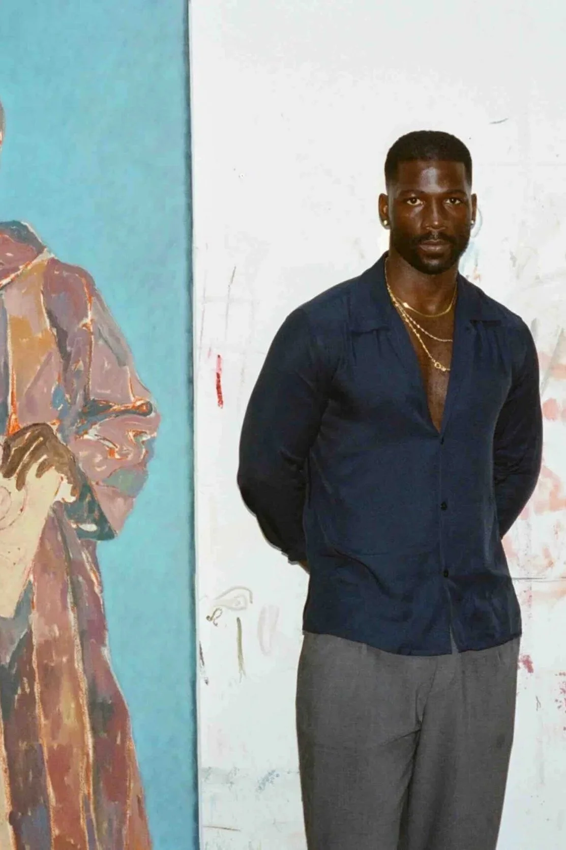 A man with short black hair and a dark complexion wearing a navy blue shirt with the top button undone, gray pants, gold necklaces, and earrings, stands in front of a white wall with a partial painting of a person wearing glasses and a cap on his left.
