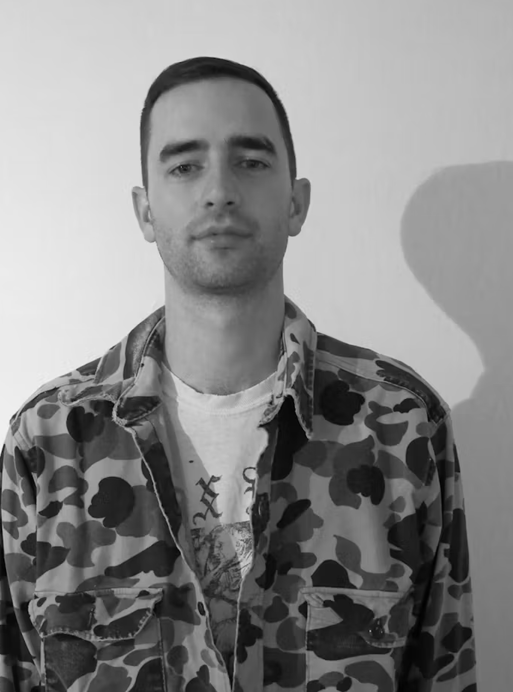 Black and white portrait of a young man with short dark hair, wearing a camouflage-patterned jacket over a graphic t-shirt, standing against a plain wall with a shadow cast on it.