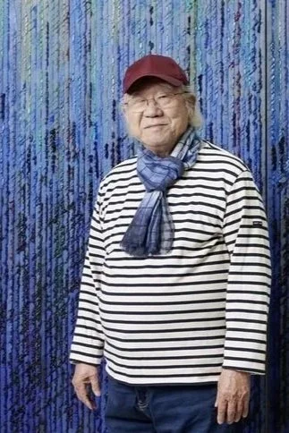 Older woman wearing a red cap, striped shirt, blue scarf, and glasses standing in front of a blue textured background.