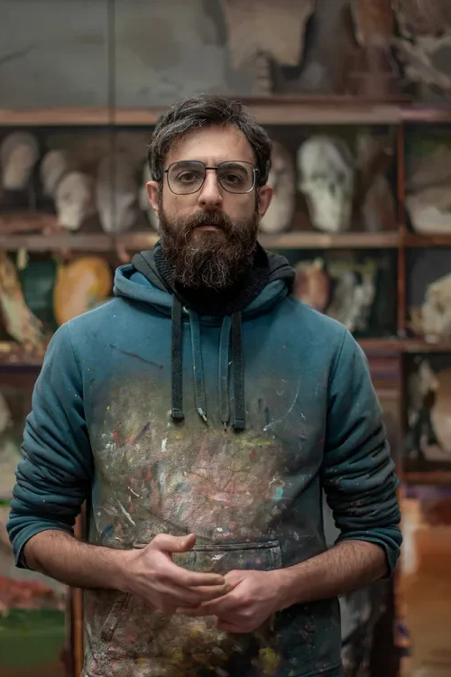 A man with glasses and a beard wearing a paint-splattered hoodie standing in front of shelves with animal skulls.